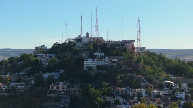 Panoramic view of communication towers and antennas crowning a hill in Durango, Mexico. White residential buildings cascade down the slopes surrounded by lush green trees under clear blue sky.