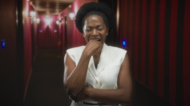 Black woman biting knuckle with hand to mouth gesture in a dim red corridor building, eyes closed, wearing white sleeveless top; pensive introspection.