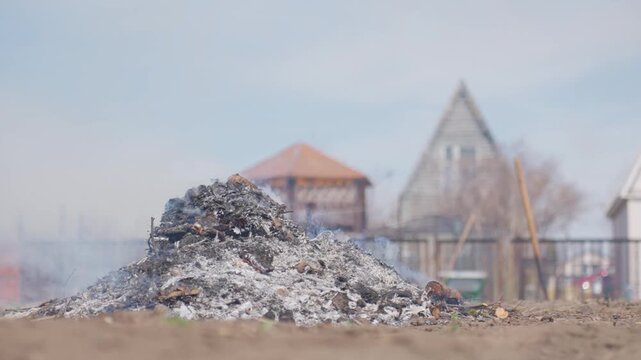 Ash Pile In Foreground Near Houses, Smoldering Heap Of Burned Debris On Vacant Lot, Blurred Suburban Rooftops And Pale Sky In Background, Thin Smoke Rising From Charred Wood And Scattered Embers, Mood