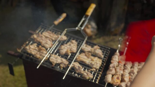 Asian Arm Basting Pork Skewers On Charcoal Grill With Red Sauce Bottle, Visible Blue Shirt Sleeve And Marinade Brush Action, Family Cookout Energy With Smoke, Sizzle And Communal Summer Atmosphere