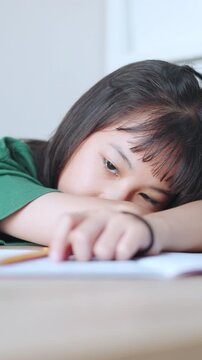 Vertical video format showing a close up and selective focus of a demotivated Asian kid girl resting her head on a desk. She looks lethargic and overworked by too much homework in bright daylight.