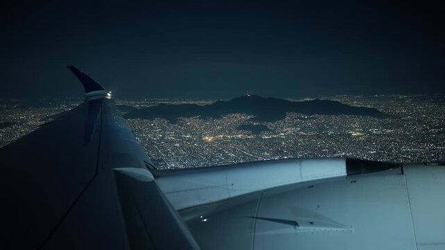 Airplane coming in for landing with view from porthole window showing vast city lights below. Aircraft wing and engine visible against dark sky with illuminated urban sprawl and mountain silhouettes.