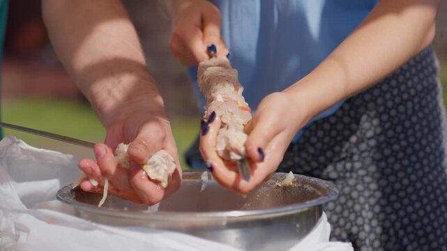 People Work Together Outdoors. Closeup Of Hands Threading Meat Onto Skewers During Outdoor Gathering. Two Individuals Coordinate To Prepare Skewered Meat In Backyard Setting For Summer Grilling