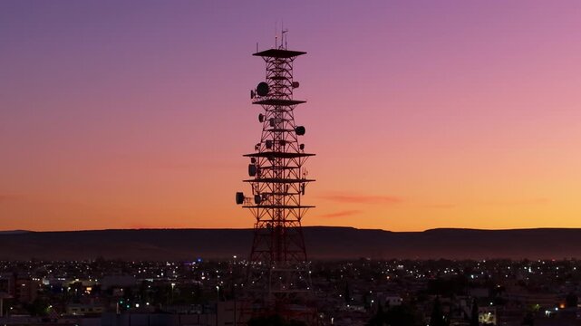 Durango, Mexico cityscape at twilight featuring a towering telecommunications antenna with multiple satellite dishes silhouetted against a stunning purple and orange gradient sunset sky.