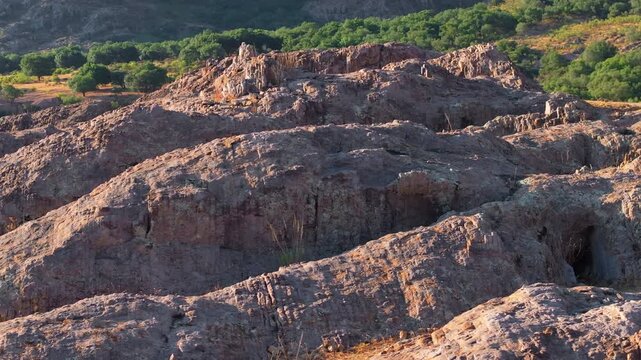 Rugged rocky landscape in Sierra Madre Occidental mountains near Durango, Mexico bathed in warm golden sunset light. Layered red and brown rock formations with caves and green forest vegetation.