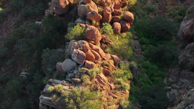 Aerial drone footage captures dramatic orange-red boulder formations scattered along a steep mountain ridge bathed in warm golden hour sunlight, surrounded by lush green vegetation and desert plants.