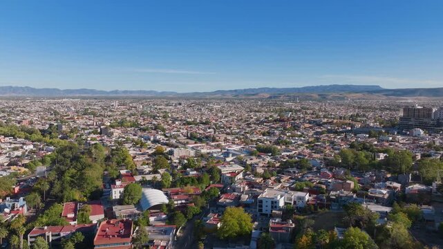 Aerial panoramic view of Durango Mexico cityscape featuring residential neighborhoods with red rooftops and green trees. Sierra Madre mountains rise in the background under clear blue sky.