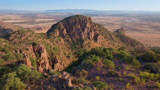 Aerial drone footage captures dramatic rocky mountain peak with rugged red cliff faces rising above arid desert plains. Golden hour sunlight illuminates sparse green vegetation and vast horizon.
