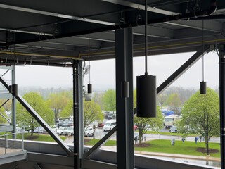 Obraz premium Modern industrial ceiling structure with exposed pipes, speakers, and beams overlooking a rainy parking lot with cars, green trees, and people walking on an overcast day.