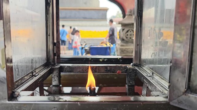 Burning candle flame inside metal lantern shrine with blurred people walking in temple background. Religious offering light in Asian spiritual sanctuary