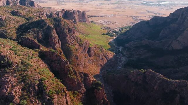 Breathtaking aerial drone footage captures rugged desert mountain terrain with dramatic red rock cliff formations, sparse green vegetation covering steep slopes.