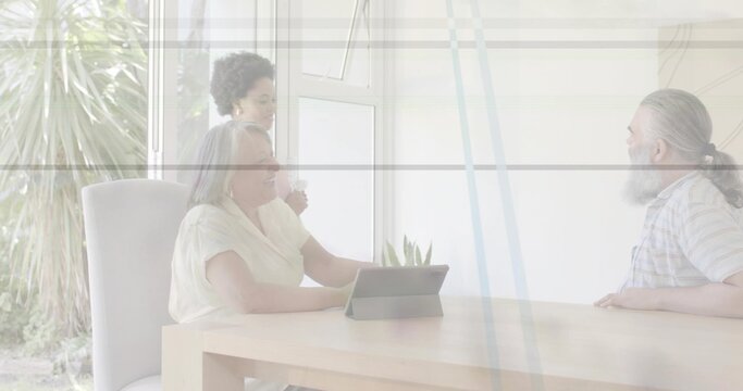 Talking three adults around wooden dining table in sunlit room, tablet visible man in checked shirt