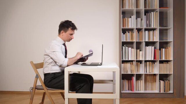 Professional man working diligently from his home office, focusing on reviewing documents and using a laptop at his desk, with a full bookshelf creating a studious background