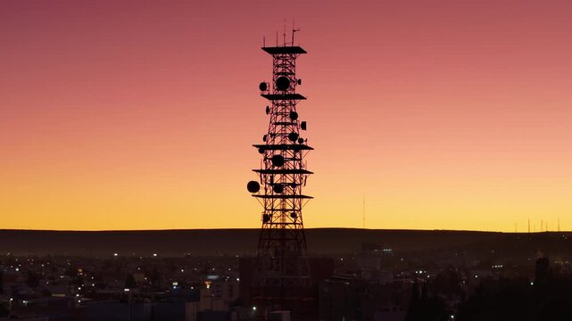 Tall communication tower with multiple satellite dishes and antennas silhouetted against stunning gradient sunset sky in Durango, Mexico. City lights twinkle below as dusk settles over urban.