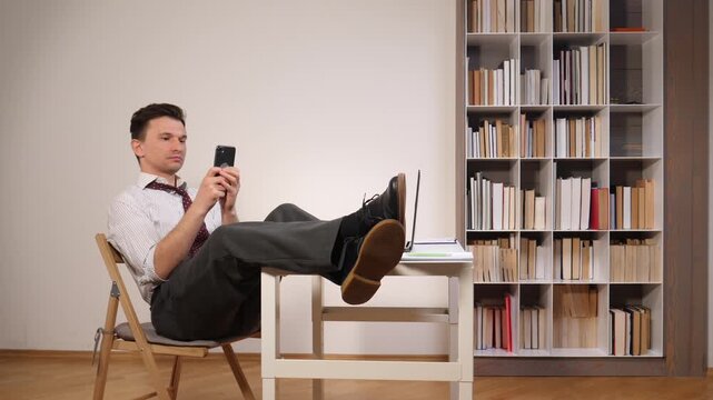 Businessman in shirt and tie with feet on desk, scrolling smartphone in a home office distracted, procrastinating and unproductive while working remotely, relaxed and disengaged