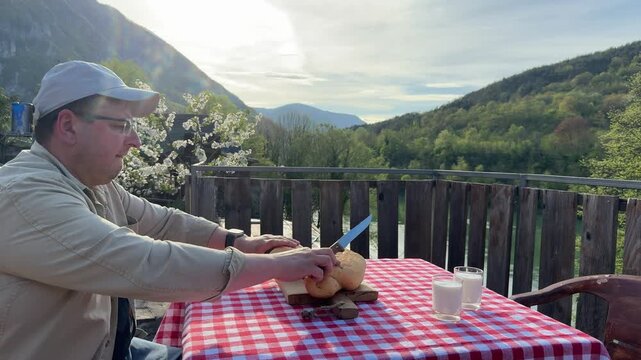 Man slicing fresh bread on wooden board at outdoor terrace with scenic mountain view in warm sunlight. Cooking outdoors and countryside lifestyle. Cozy countryside lifestyle series &mdash; outdoor cooking