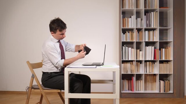 Man in an office setting looking critically at his empty wallet, reflecting on financial difficulties and struggling with money management or personal debt while working from his desk at home