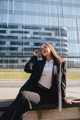Fototapeta premium Beautiful Business Woman Sitting Outdoors Near Modern Office Building Wearing Black Blazer And Glasses. Corporate Lifestyle Concept Female Professional Urban