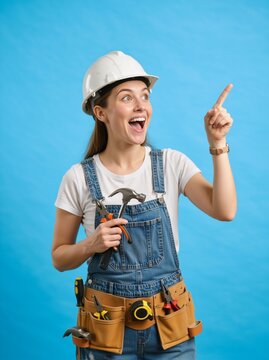 Vertical portrait of an excited female construction worker pointing up. Young handywoman in a hard hat and overalls holding tools on a blue background