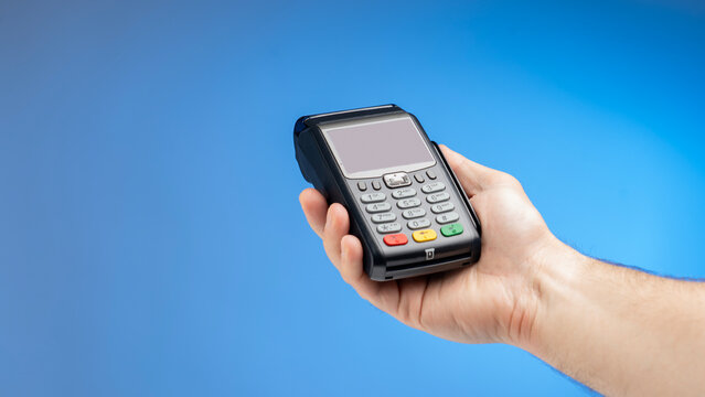 Hand holds a black payment terminal. Blue background fills the scene. Terminal has a screen and keypad. Buttons include red, green, and yellow. Hand is steady and focused