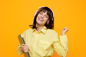A cheerful student wearing a yellow shirt and pink headphones smiles against a bright orange backdrop, holding a notebook as she enjoys music and study in a lively scene. © SHOTPRIME STUDIO