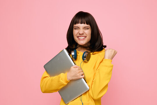 A cheerful student stands against a pink backdrop, holding a laptop with headphones around the neck. Dressed in a bright yellow hoodie, the scene radiates creativity, confidence, and friendly energy.