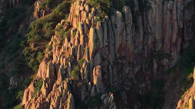 Aerial view of dramatic granite rock formations and towering stone pillars bathed in warm golden hour sunlight with lush green vegetation clinging to the rugged mountainside in Mexico.