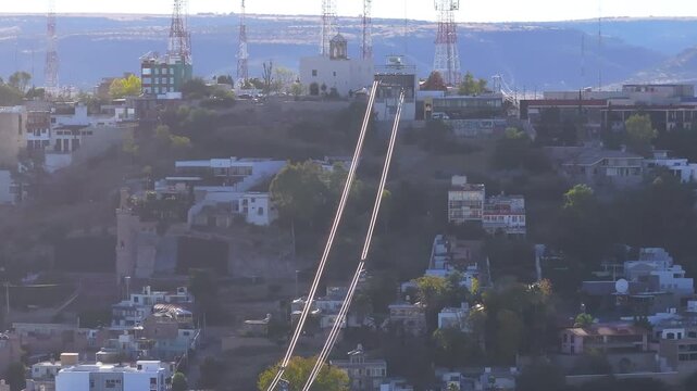 Cable car gondola travels along cables through hillside residential neighborhood in Durango, Mexico. Communication towers and white buildings dot landscape as warm sunset light illuminates the scene.