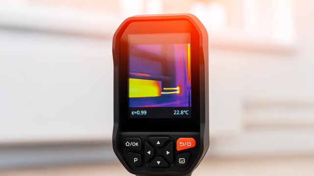 Close-up shows a handheld thermal camera. Its screen displays temperature readings with color gradients. The device has buttons labeled OK and P. Background is softly blurred indoor scene