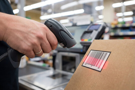 A handheld barcode scanner scans a cardboard box at a retail checkout counter. The composition focuses on checkout workflow with the reader aligned toward the barcode label.
