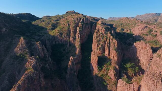 Aerial drone footage captures stunning red rock canyon formations with dramatic vertical cliffs and spires bathed in warm golden hour sunlight against a clear blue sky in rugged mountain terrain.