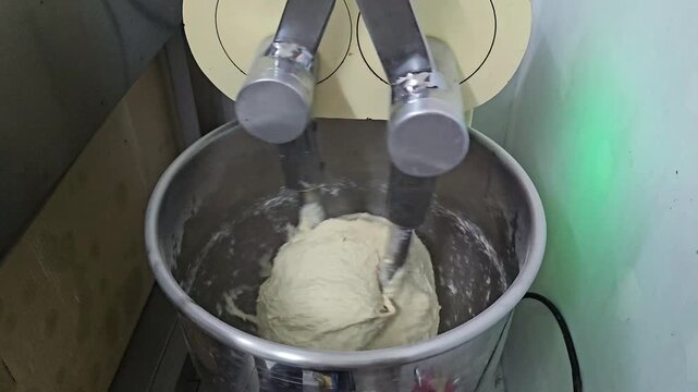 Industrial bakery mixer kneading dough in metal bowl with rotating arms, top view of food processing machine working with elastic dough mass, continuous mixing process in professional kitchen equipmen