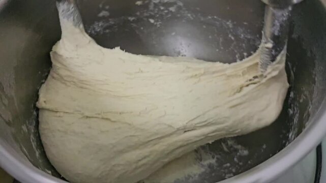 Industrial dough mixing process in metal bowl with rotating hooks kneading elastic mass, close up of bread dough preparation in bakery equipment, continuous motion forming smooth textured mixture