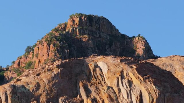 Majestic rocky mountain peak with rugged cliff faces and sparse green vegetation rises against clear blue sky as golden dry grass sways in foreground during warm sunset light in arid landscape.