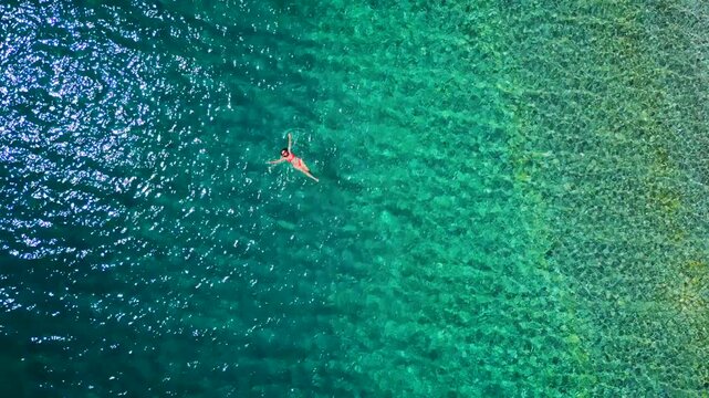 Woman in a pink swimsuit swims on her back in the clear sea, top down view