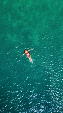 Woman in a red swimsuit swims on her back in the clear sea, top down view