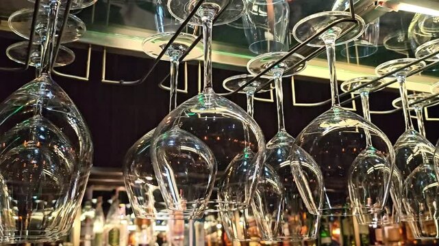 Hanging inverted wine glasses on metal rack above bar counter with illuminated bottles and reflections, close up angle capturing glassware storage and nightlife restaurant interior ambiance