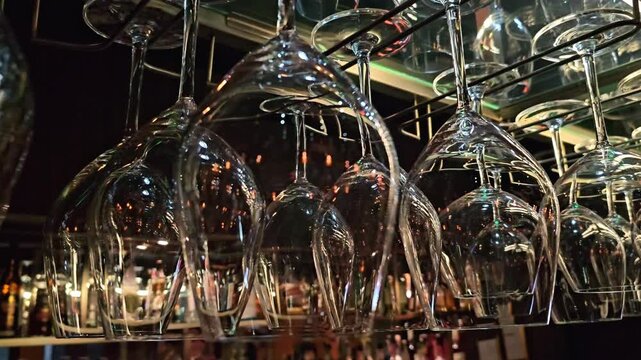 Rows of upside down wine glasses hanging on rack above bar counter with bottles in background, close up view through glassware with reflections and warm ambient lighting in restaurant interior