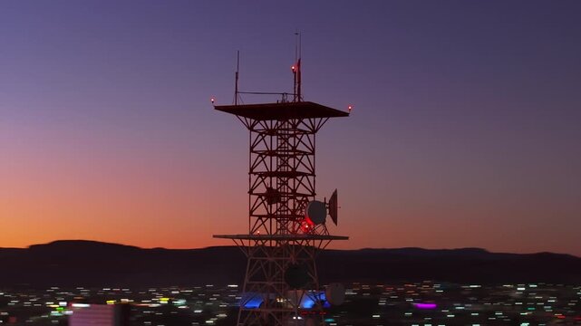 Aerial view of telecommunications tower with satellite dishes and antennas silhouetted against vibrant sunset sky in Durango, Mexico. City lights twinkle below as purple and orange hues blend.