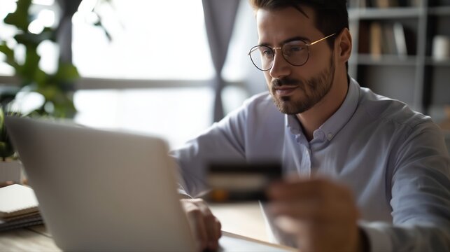 A man sits at a modern home office desk using a laptop and holding a bank card. He appears focused on making an online purchase, surrounded by plants and notebooks.