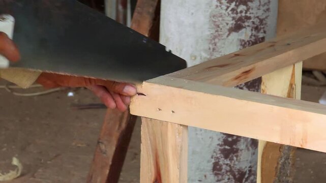 Carpenter cutting through a wooden plank with a manual hand saw in a rustic outdoor workshop environment High Quality Footage