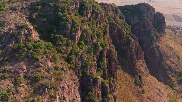 Drone footage captures rugged terrain of Sierra Madre Occidental in Durango, Mexico. Rocky cliffs covered with lush green vegetation rise above arid farmland and desert plains during golden hour.
