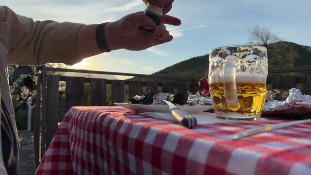 Close-up of adding seasoning to food during an outdoor picnic dinner on a terrace with scenic mountain view. Warm sunset light, lens flare, and rustic countryside atmosphere. Concept of cooking, outdo