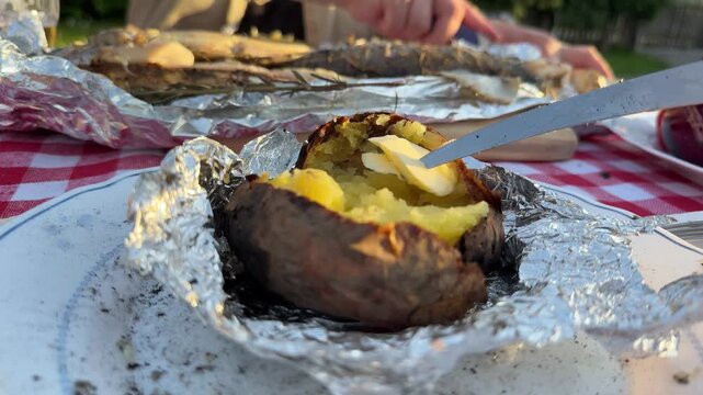POV close-up shot of hands cutting baked potato wrapped in foil at an outdoor picnic table. Traditional rustic meal with grilled fish, beer, and homemade food. Warm sunset light and countryside atmos