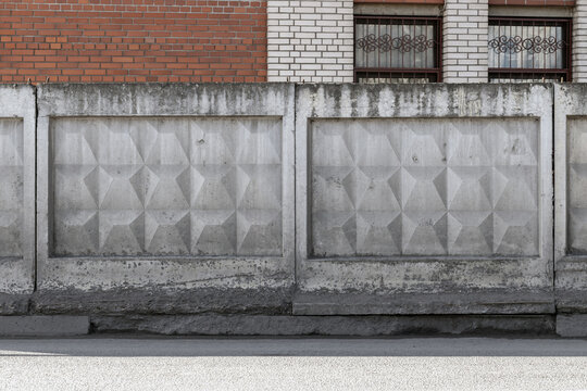 Brick wall of an industrial building is behind a weathered concrete fence featuring geometric relief panels and repeating textured pattern. Urban architectural facade shows stains and decay
