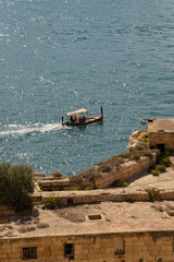 Naklejka premium Vertical view of a traditional Maltese dghajsa boat sailing near historic bastions. Sunny Mediterranean sea in Valletta, Malta.