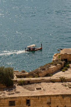 Vertical view of a traditional Maltese dghajsa boat sailing near historic bastions. Sunny Mediterranean sea in Valletta, Malta.