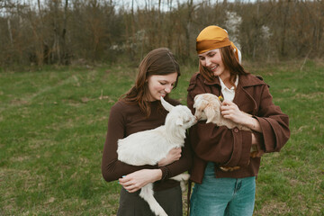 Fototapeta premium Lifestyle scene with two women outdoors holding a kid goat, sharing a gentle moment. Natural light, grassy field, casual wear, connection with animals, promoting wellness and friendship.
