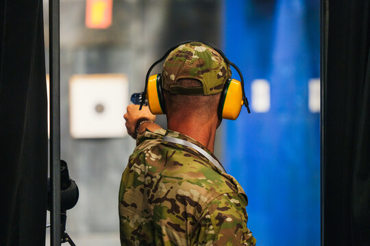 Man in a camouflage uniform stands with his back to the camera in a shooting range and holds a pistol, aiming at a target in the distance.