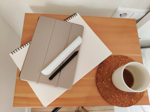 Tablet with stylus, notebook and coffee cup on wooden bedside table. Minimalist interior workspace for study, planning or creative work. Organization of home office or desk space.